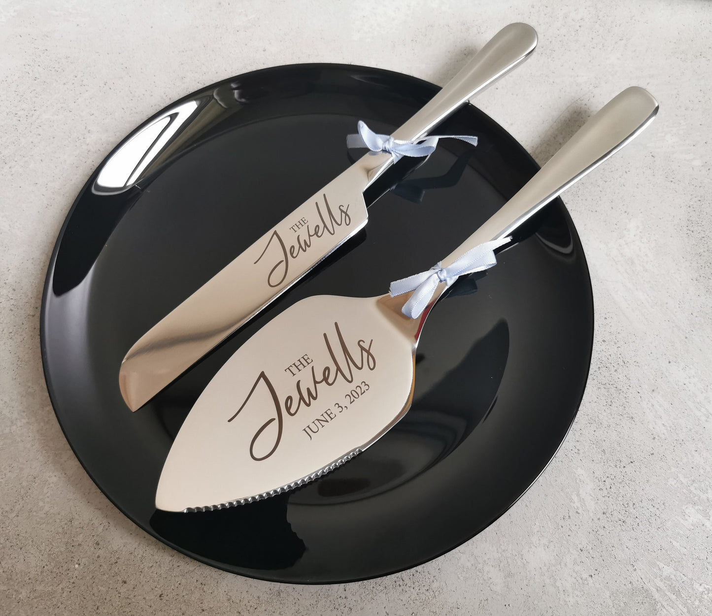 May include: Two silver cake servers with black handles. The top server is a knife and the bottom server is a spatula. Both servers are engraved with the text 'The Jewells' and the date 'June 3, 2023'.
